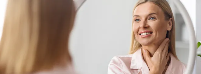 Woman smiling in mirror and gently touching neck during neck lift recovery at home